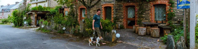 Promeneur et son chien passant devant un gîte en pierre avec terrasse arborée